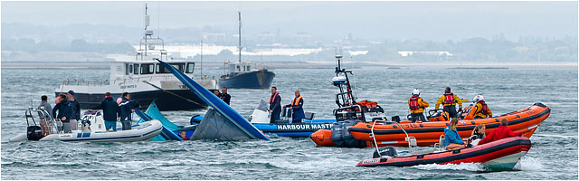 Capsized Catamaran Boat With Sailor In Water Surrounded By Rescue Teams