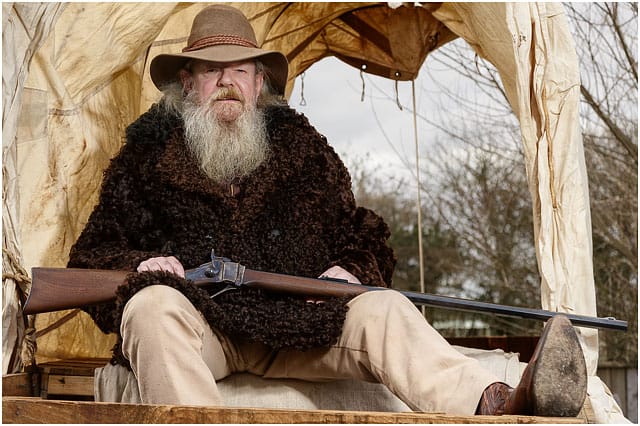 Portrait Of Man Dressed As An American Wild West Trapper Sat In Covered Wagon With A Shot Gun