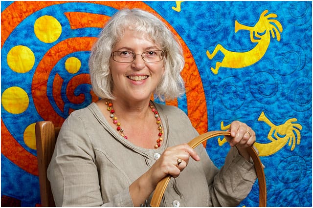 Headshot Portrait Of Textile Artist Holding Wooden Quilting Hoop In Front of Quilted Kokopelli Figures