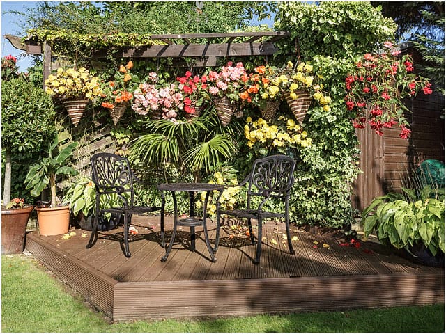 Wooden Pergola Decking Metal Table Chairs Hanging Baskets Red Yellow Pink Flowers Green Foliage Blue Sky 