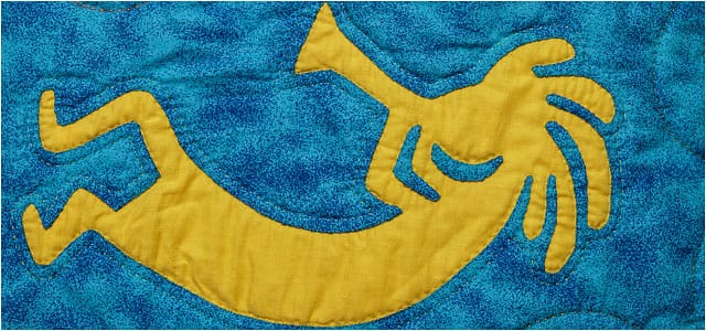 yellow quilted kokopelli figure playing musical instrument on a blue background 