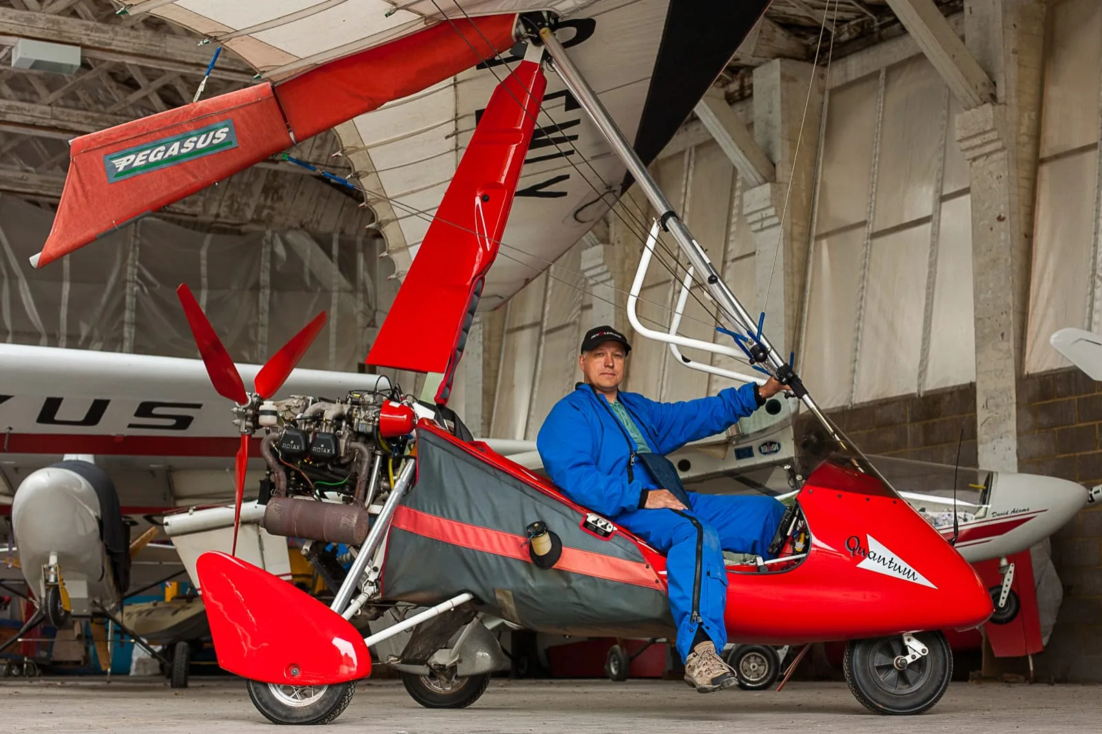 promotional portrait of middle aged man wearing blue overalls sat in the cockpit of his red microlight aircraft in the hanger