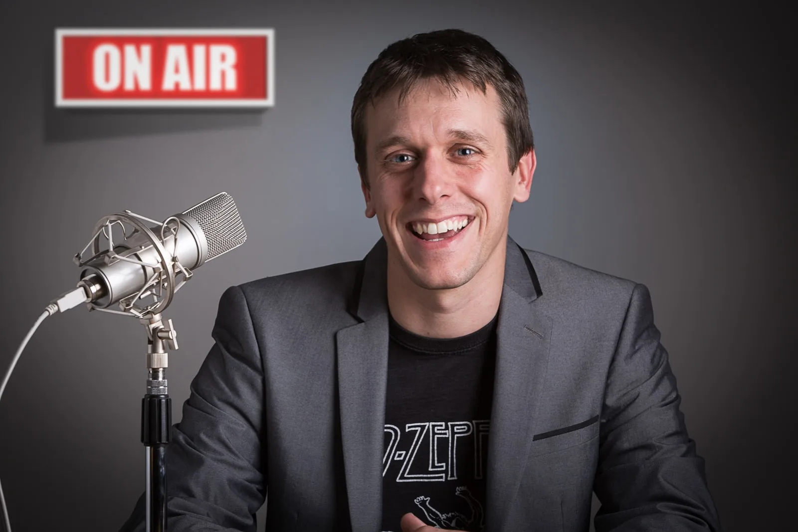 social media portrait of male audio recording artist wearing a T-shirt and business jacket with microphone and 'on air' sign in the background