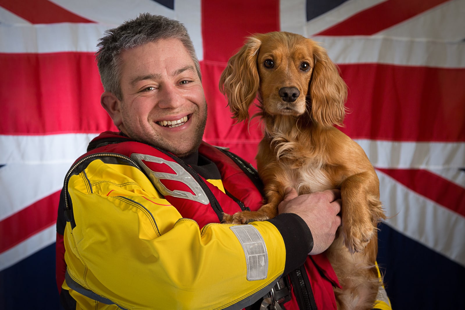 portrait of male Royal National Lifeboat Institution volunteer holding spaniel dog with British flag as a background