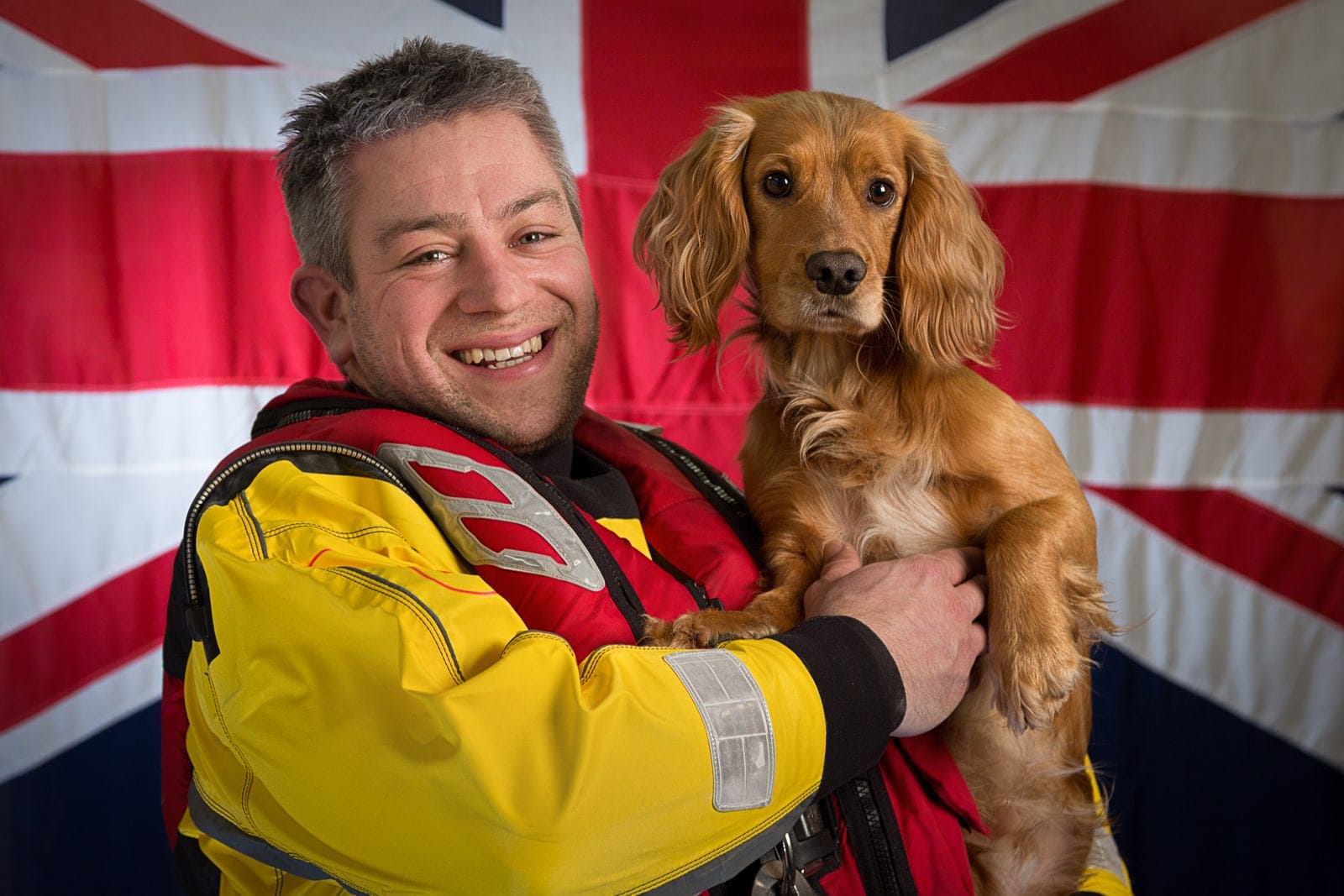 portrait of male Royal National Lifeboat Institution volunteer holding spaniel dog with British flag as a background