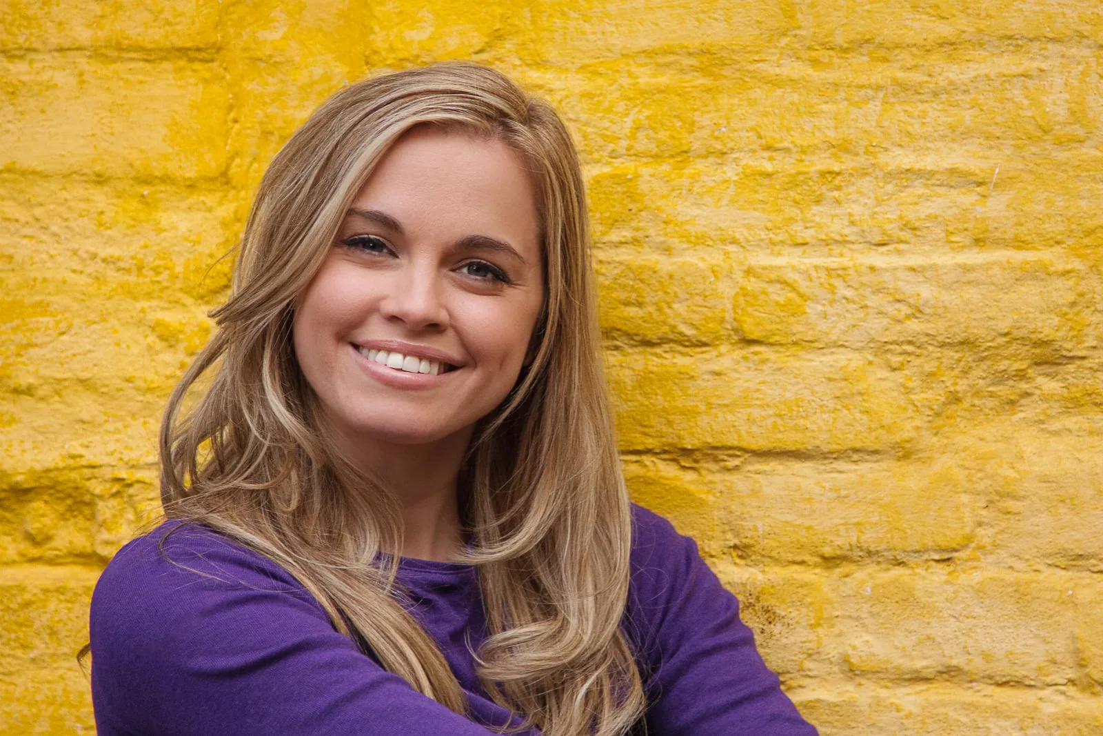 portrait of young blonde female with purple top against a bright yellow painted brick wall